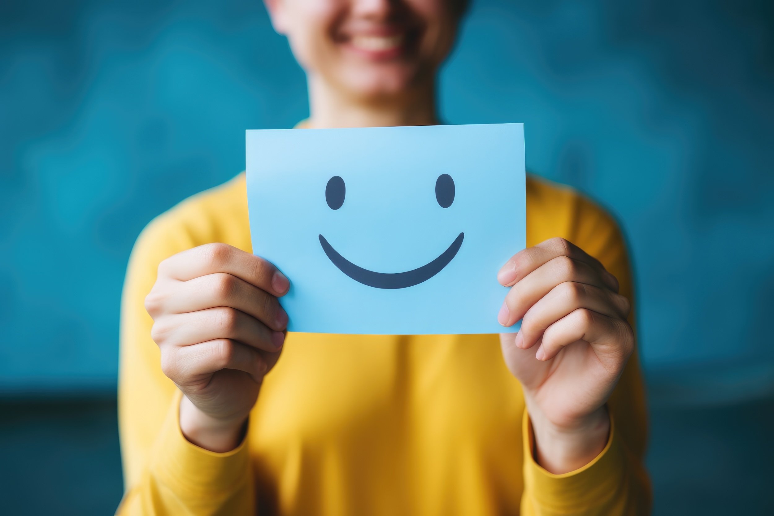 A person in a yellow shirt holding up a light blue paper with a simple smiley face drawn on it in front of their face, obscuring it. The background is a blurred blue texture, enhancing the cheerful and positive theme of the image.