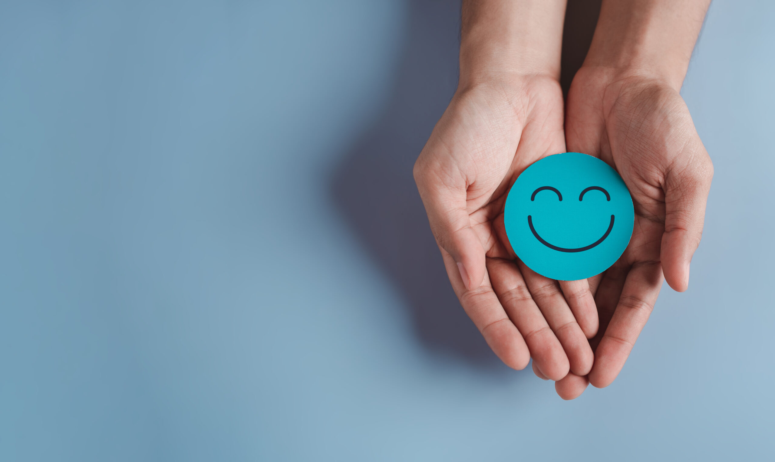 Two hands gently holding a small, round, blue smiley face. The background is a solid light blue color, providing a calm and minimalist appearance to the image.