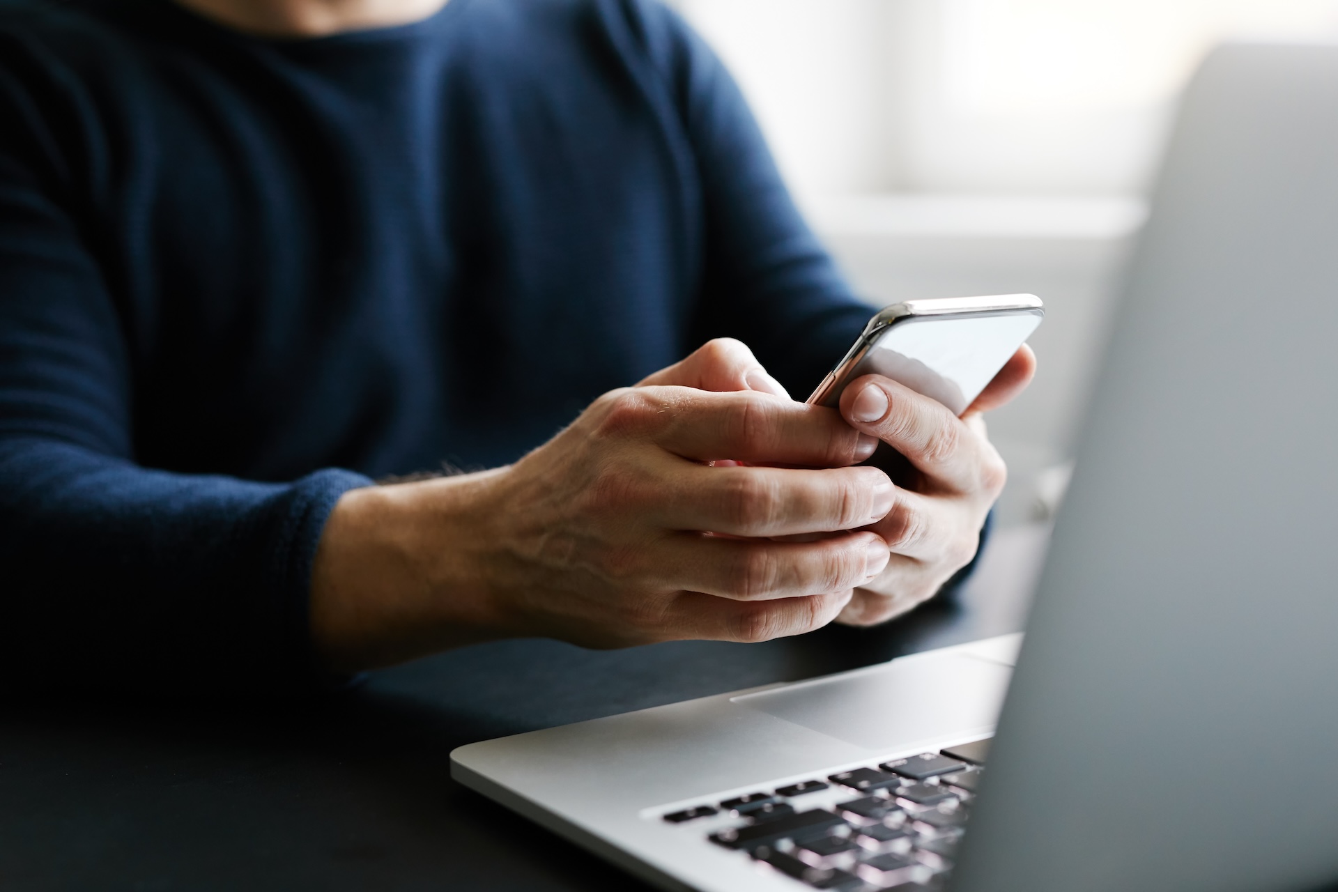 Close-up of a man's hands using a smartphone while sitting at a desk with a laptop.