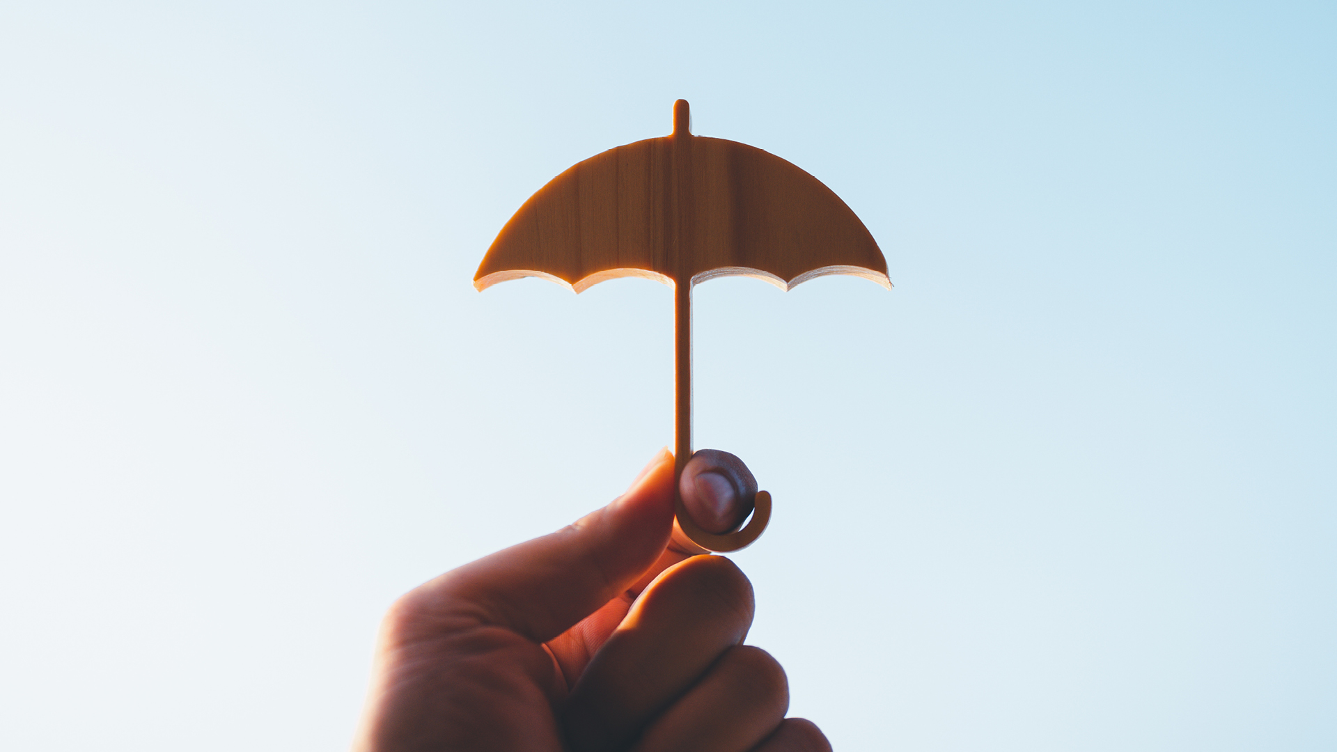 A hand holding a small wooden umbrella model against a clear blue sky, symbolizing protection and safety.