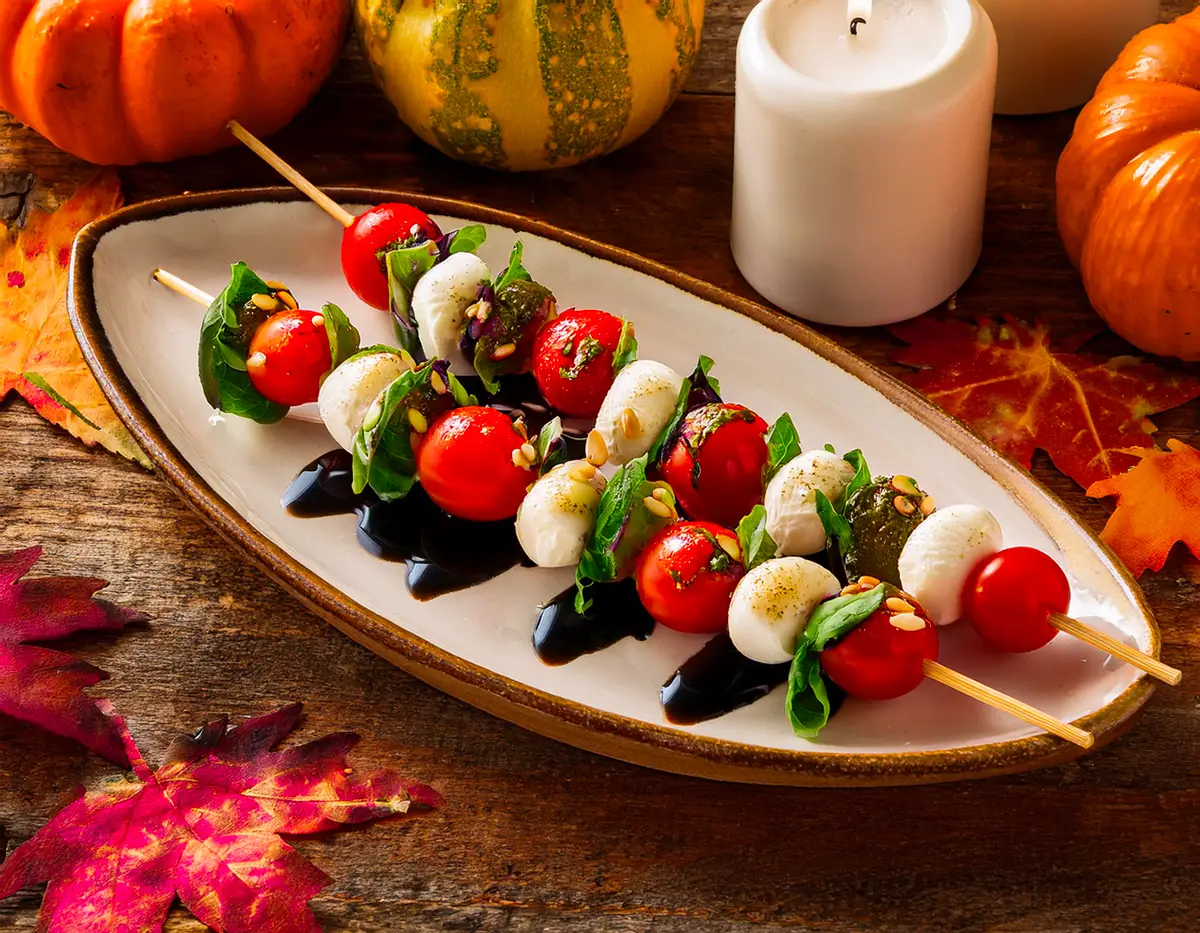 True-Thanksgiving-Recipe-Harvest-Herb-Caprese-Skewers Harvest Herb Caprese Skewers displayed on a rustic oval plate. Each skewer is stacked with cherry tomatoes, fresh mozzarella balls, and basil leaves, drizzled with balsamic glaze and sprinkled with toasted pine nuts for an appetizing presentation.