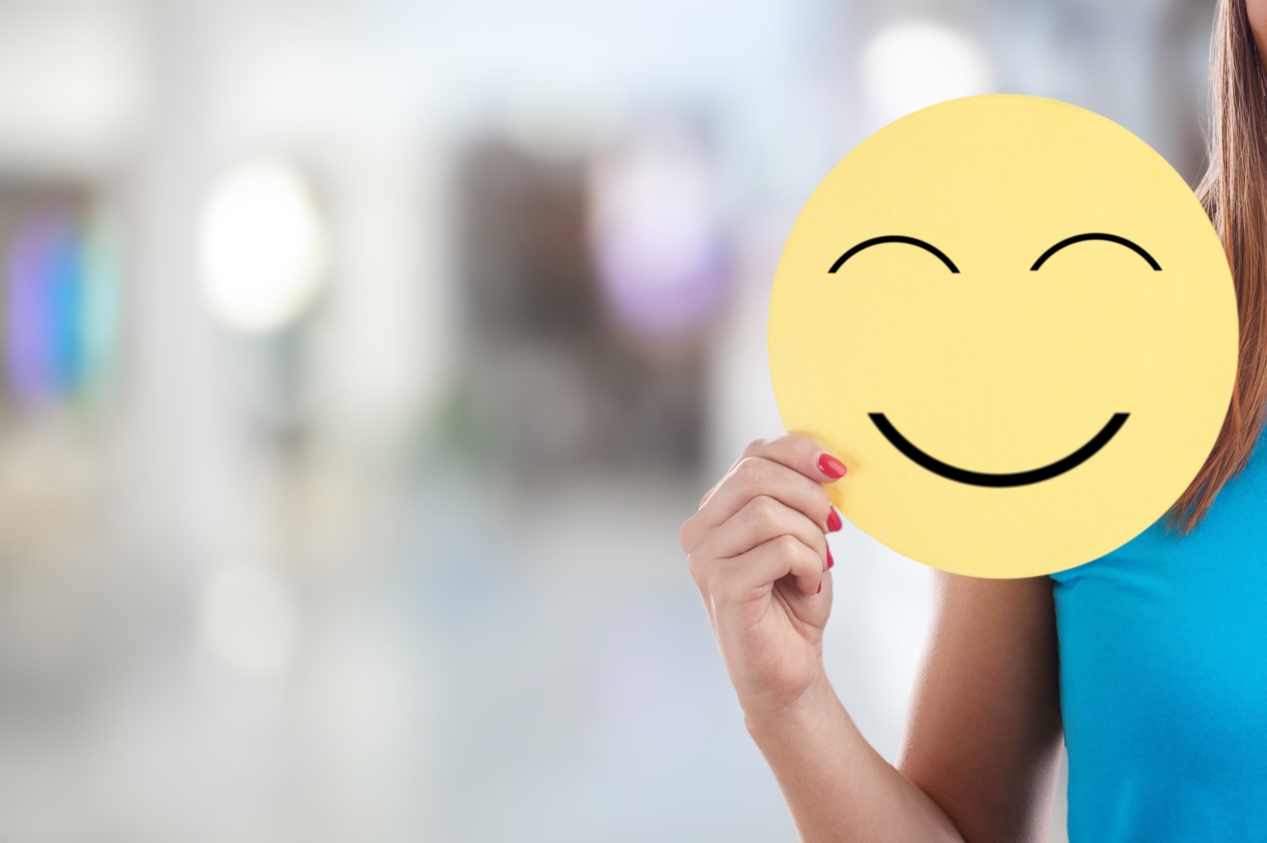 Close-up of a person holding a yellow smiling face cutout in front of their own face, symbolizing happiness and positivity in a modern office environment with a blurred background.